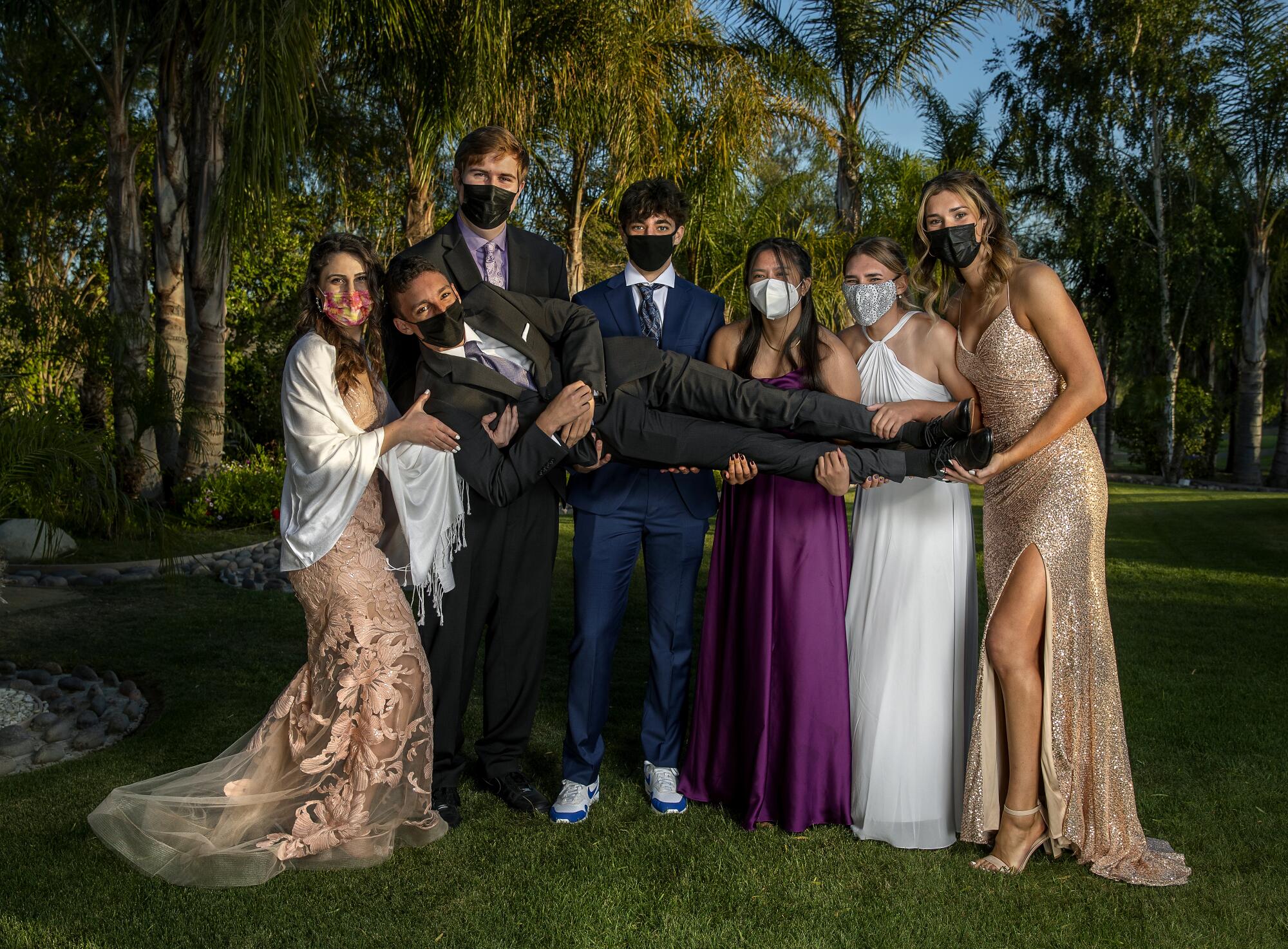 Students in prom attire pose for a photo holding one of their friends and wearing masks