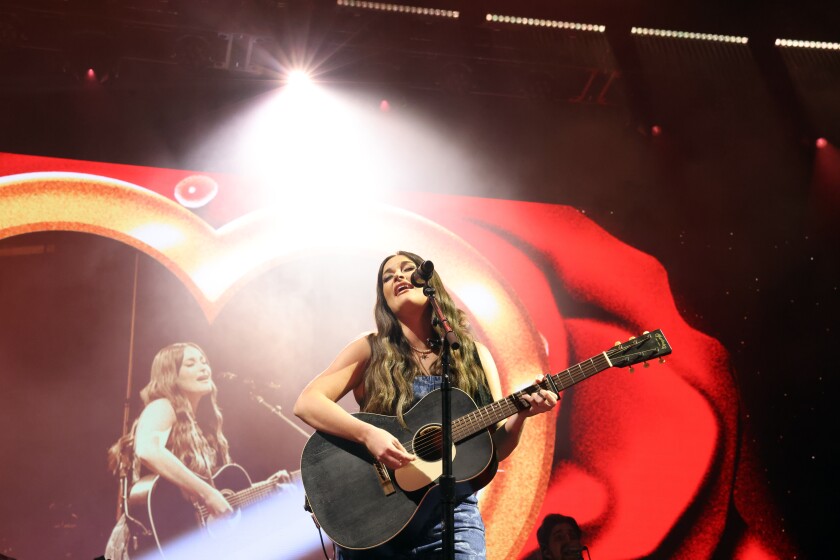 A female singer performs onstage with an acoustic guitar.