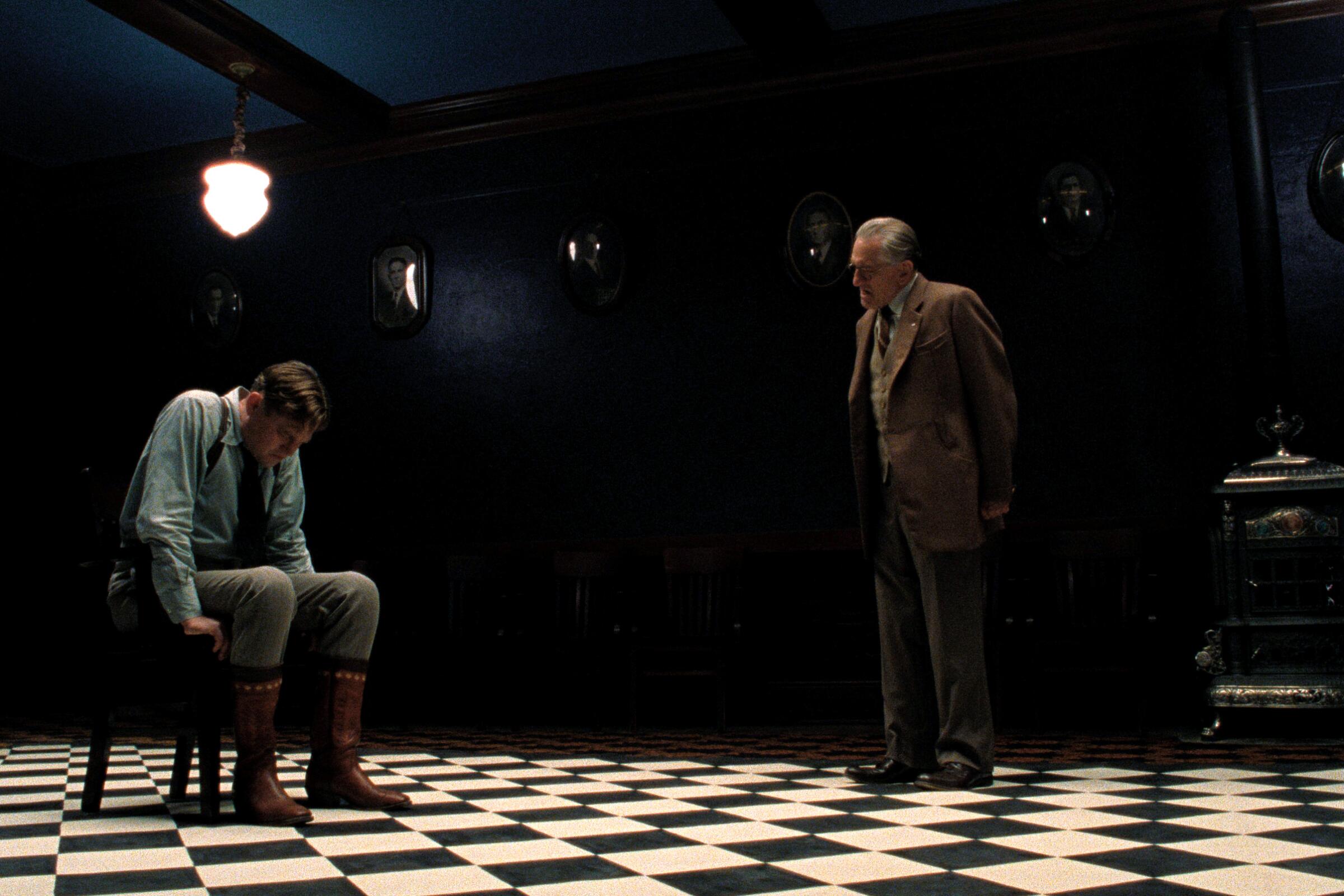 A man in a darkened room sits in a chair, head hanging down while another man lectures him in "Killers of the Flower Moon."