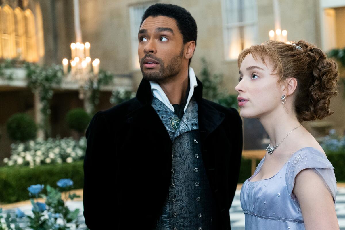 Regé-Jean Page and Phoebe Dynevor look off camera with flowers and candles behind them. They are wearing early-1800s finery.