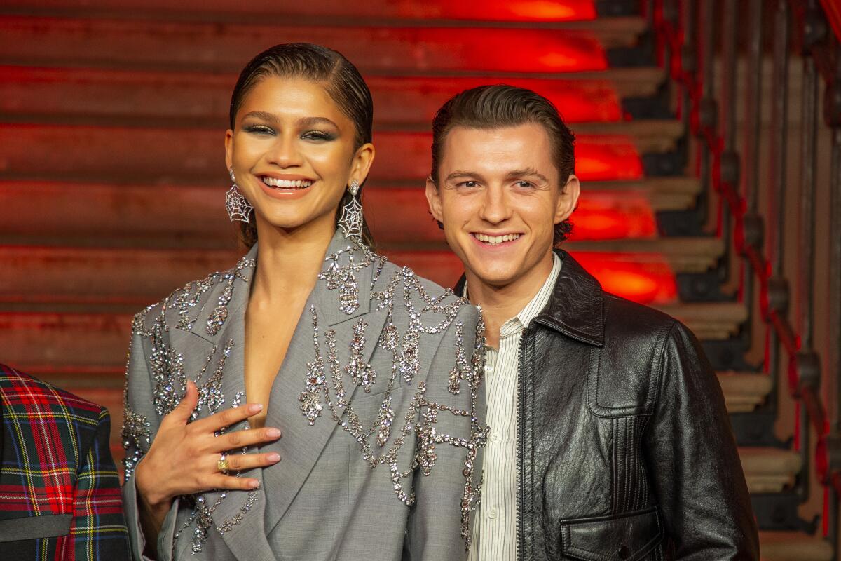 Zendaya stands in a bedazzled jacket with one hand on her chest next to Tom Holland in a leather jacket