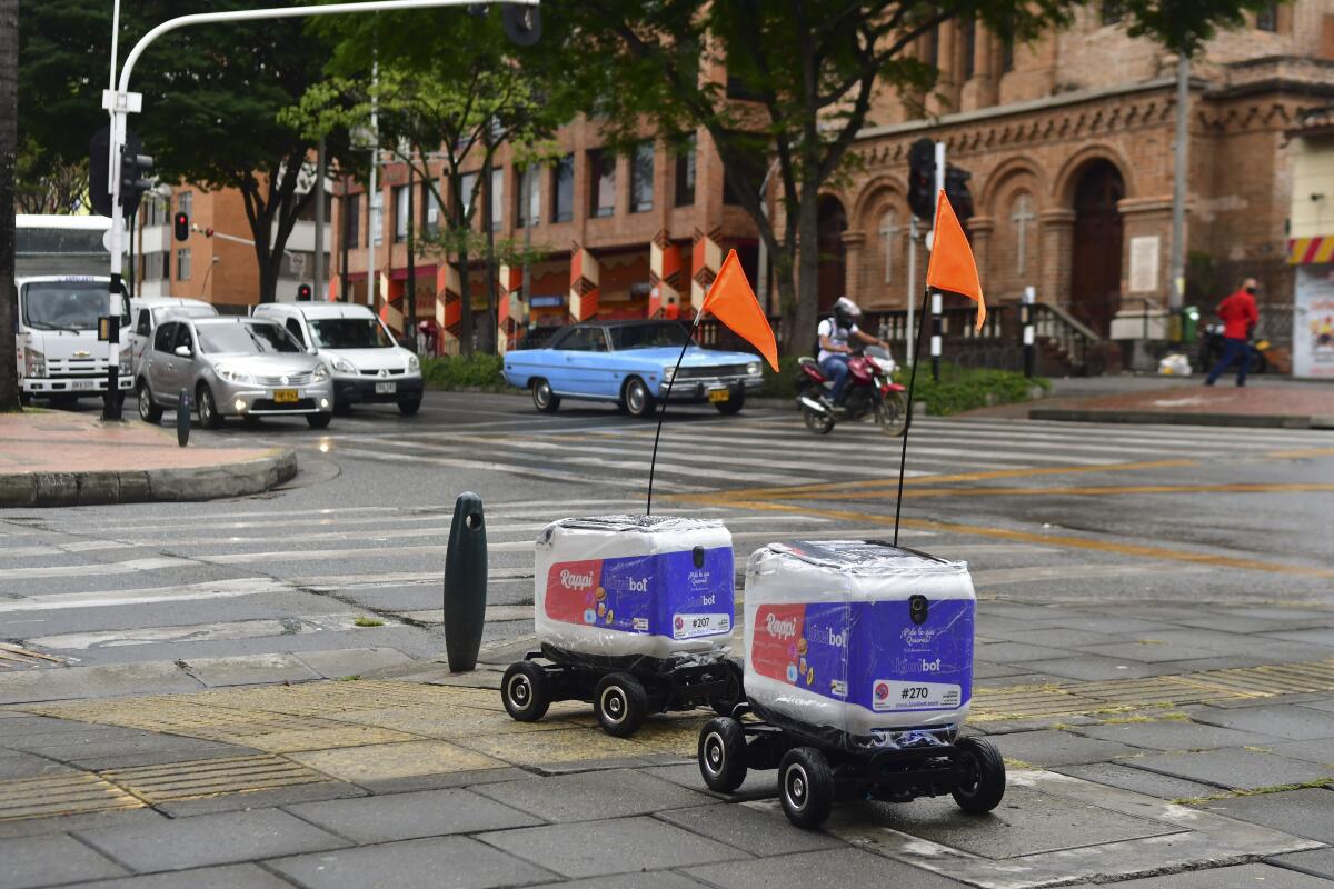 Two delivery robots, looking like little coolers on wheels with a flag attached, roll down a city street.