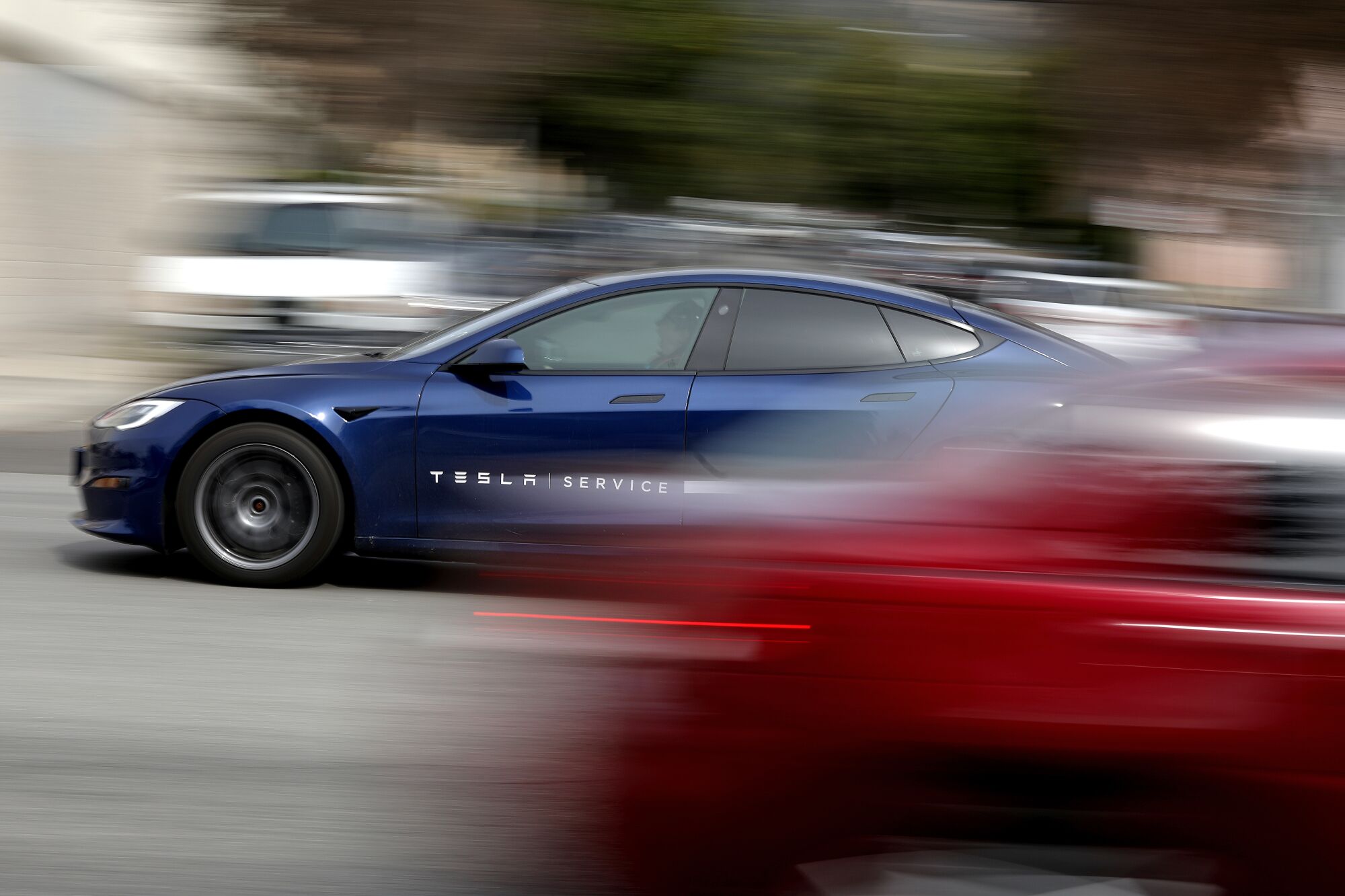 A blue car drives on a street, passing a red car, which is streaked by the panning camera.