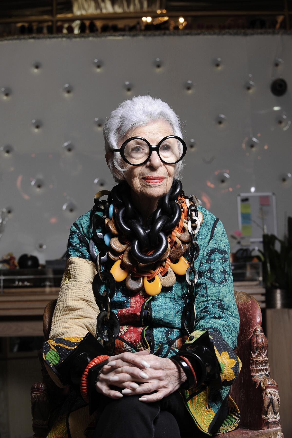 Iris Apfel, in big round glasses, a colorful ensemble and layers of necklaces, sits in a chair with her hands crossed.
