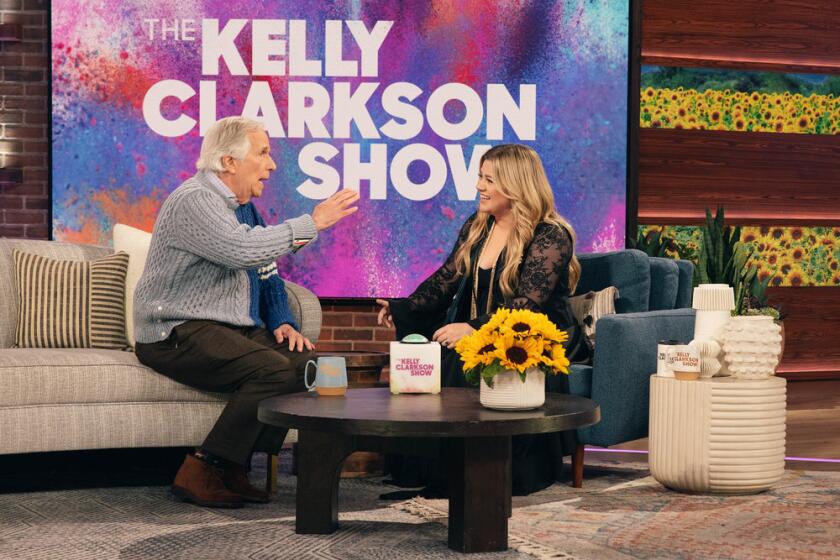 Henry Winkler sits on a couch gestures while talking to Kelly Clarkson on a talk-show set.