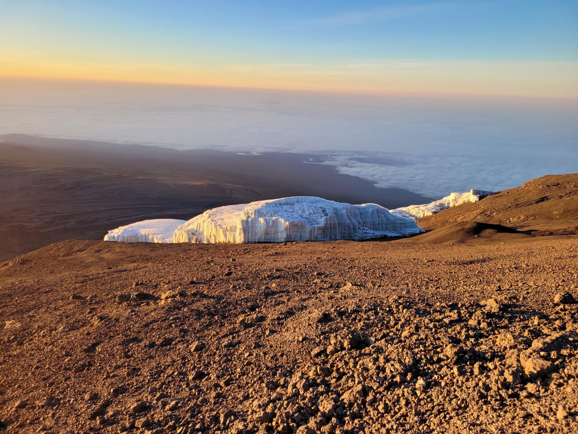 A chunk of icy rock is surrounded by brown soil with blue and orange skies overhead