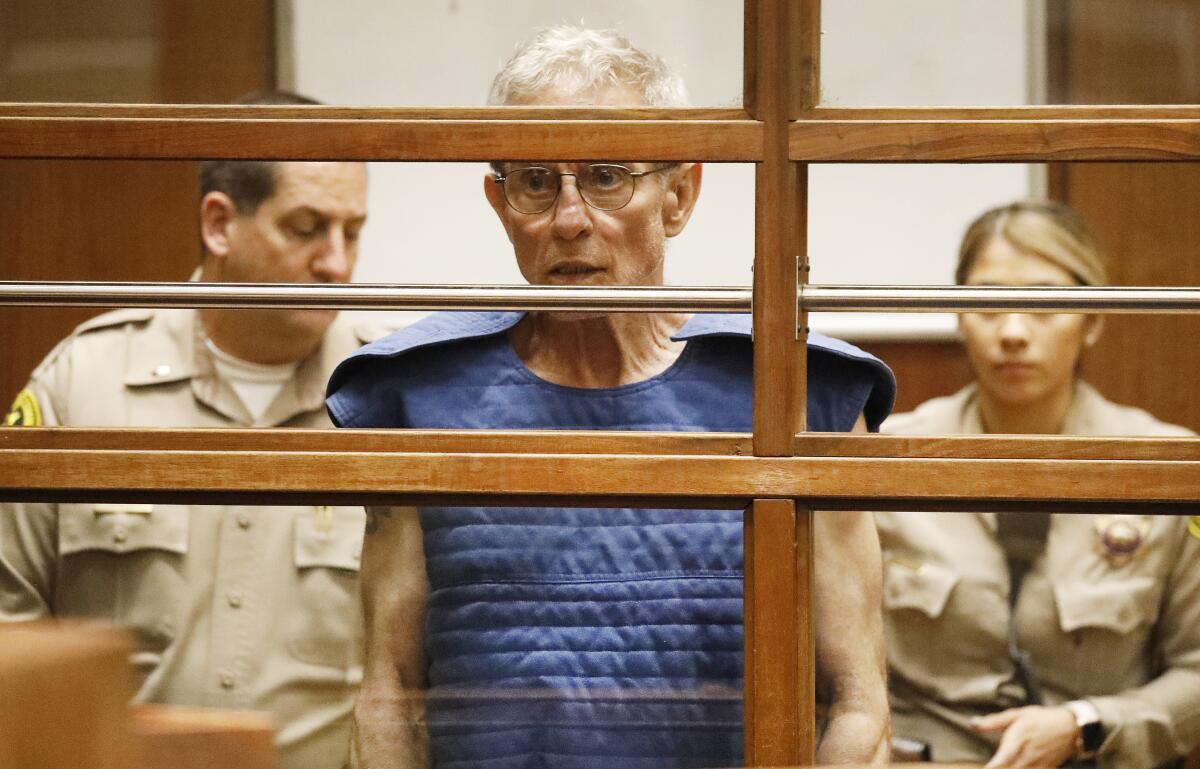 Ed Buck in a blue vest in a courtroom, with two deputies standing behind him