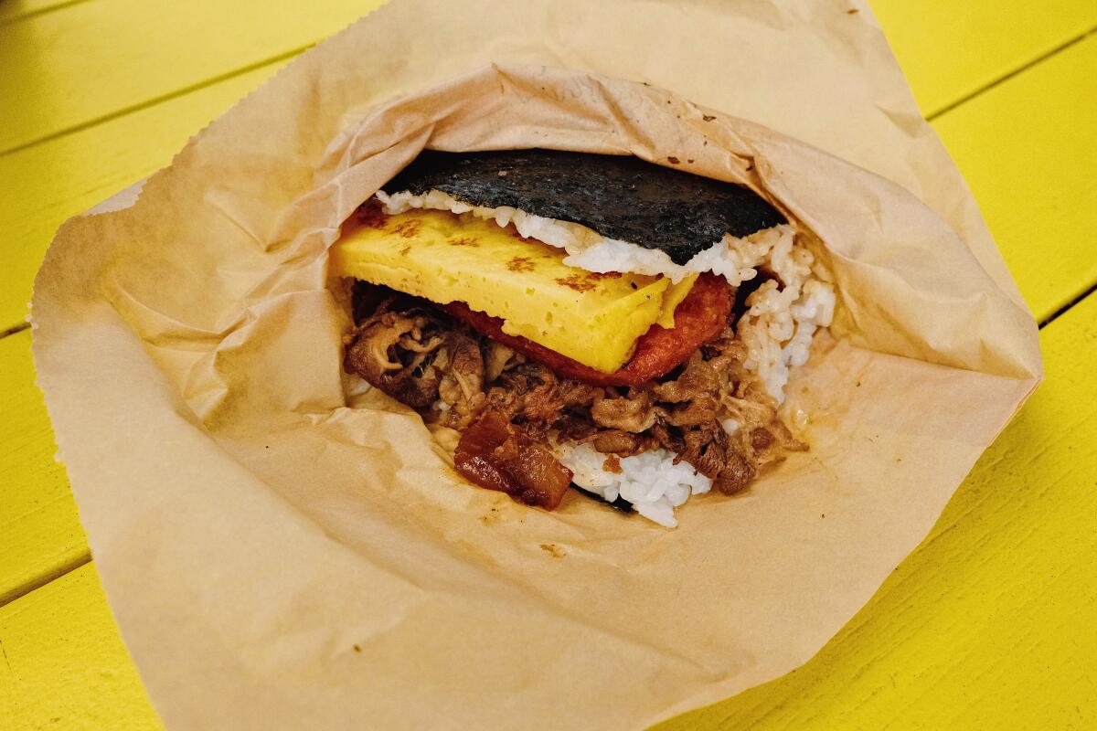 Spam musubi with sukiyaki in brown paper on a bright yellow table at Supamu restaurant in Koreatown.
