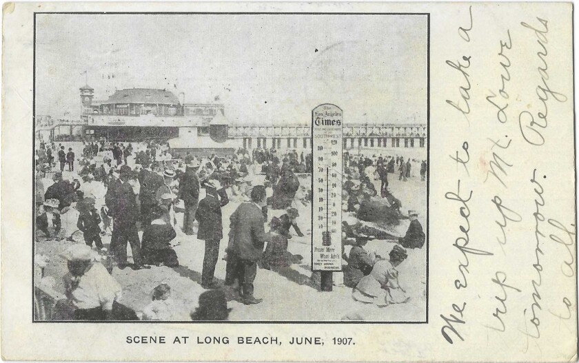 Men in suits and women in dresses and hats gather around a thermometer on the sand in Long Beach.