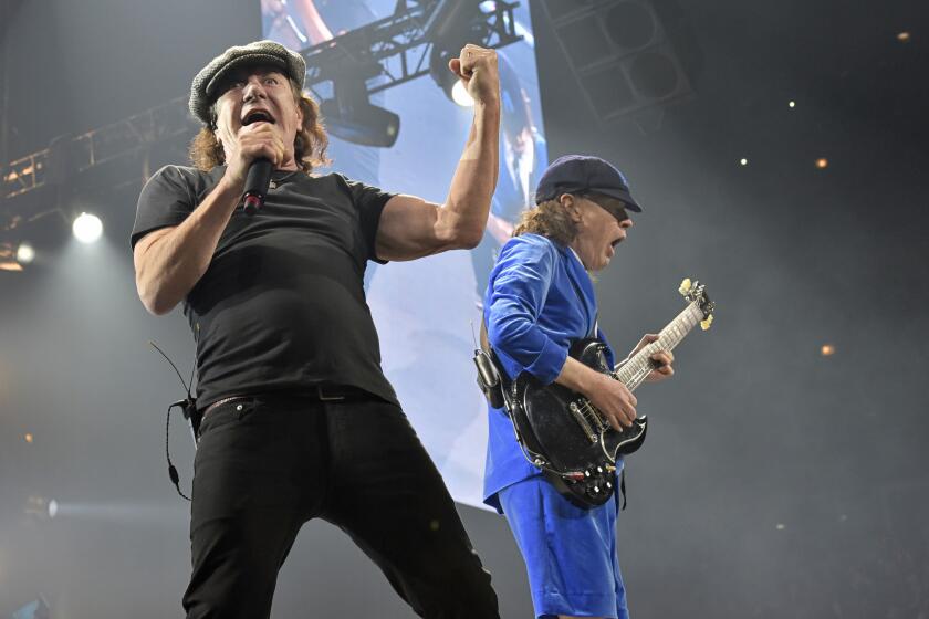 A man in black sings into a microphone while a man wearing blue plays guitar behind him