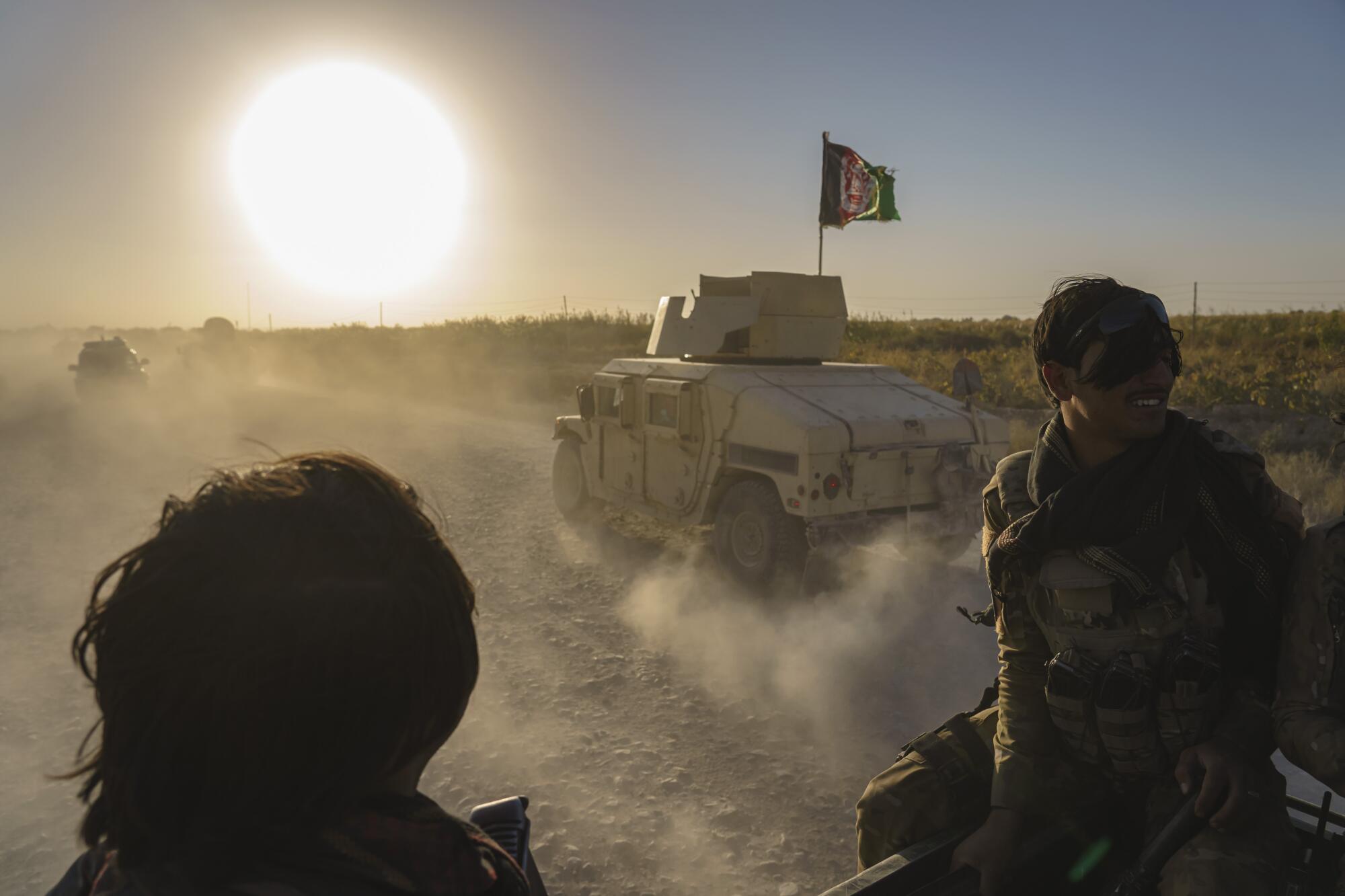  Two people are seen, dark against the sun's glare, as military vehicles kick up clouds of dust on a rocky road.