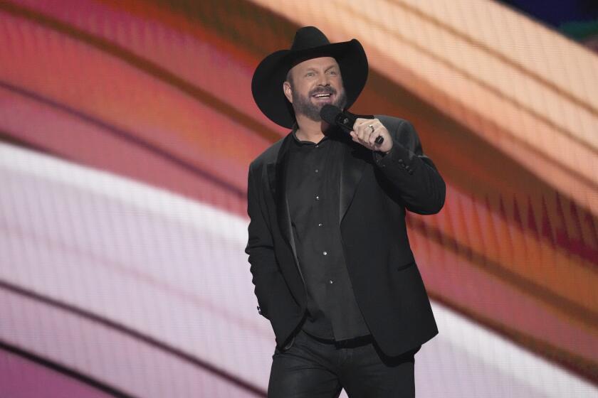 Garth Brooks stands onstage with one hand in his pocket while speaking into a mic. He wears a black cowboy hat and blazer.