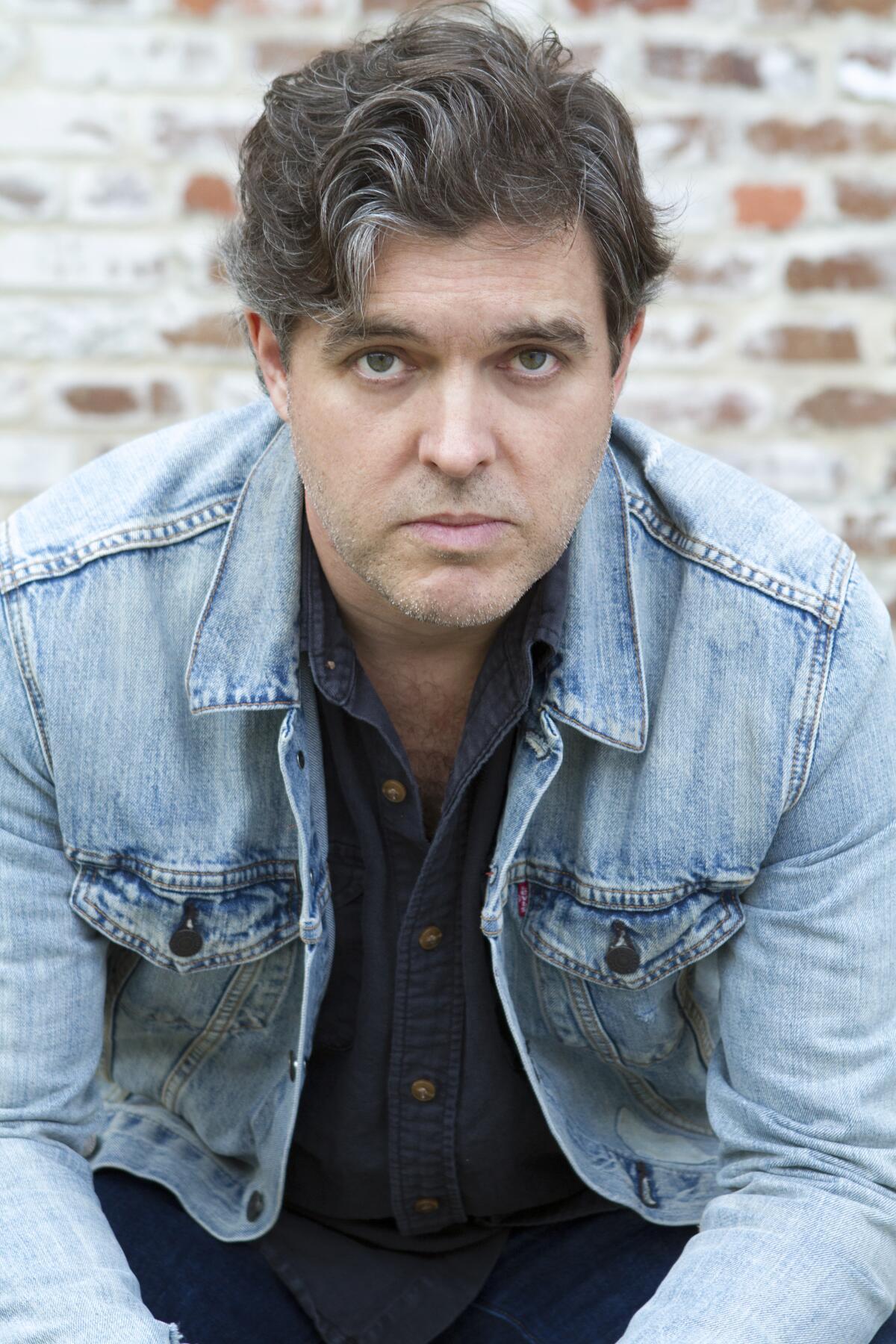 Man with wavy salt-and-pepper hair in blue shirt and denim jacket, seated in front of a brick wall.