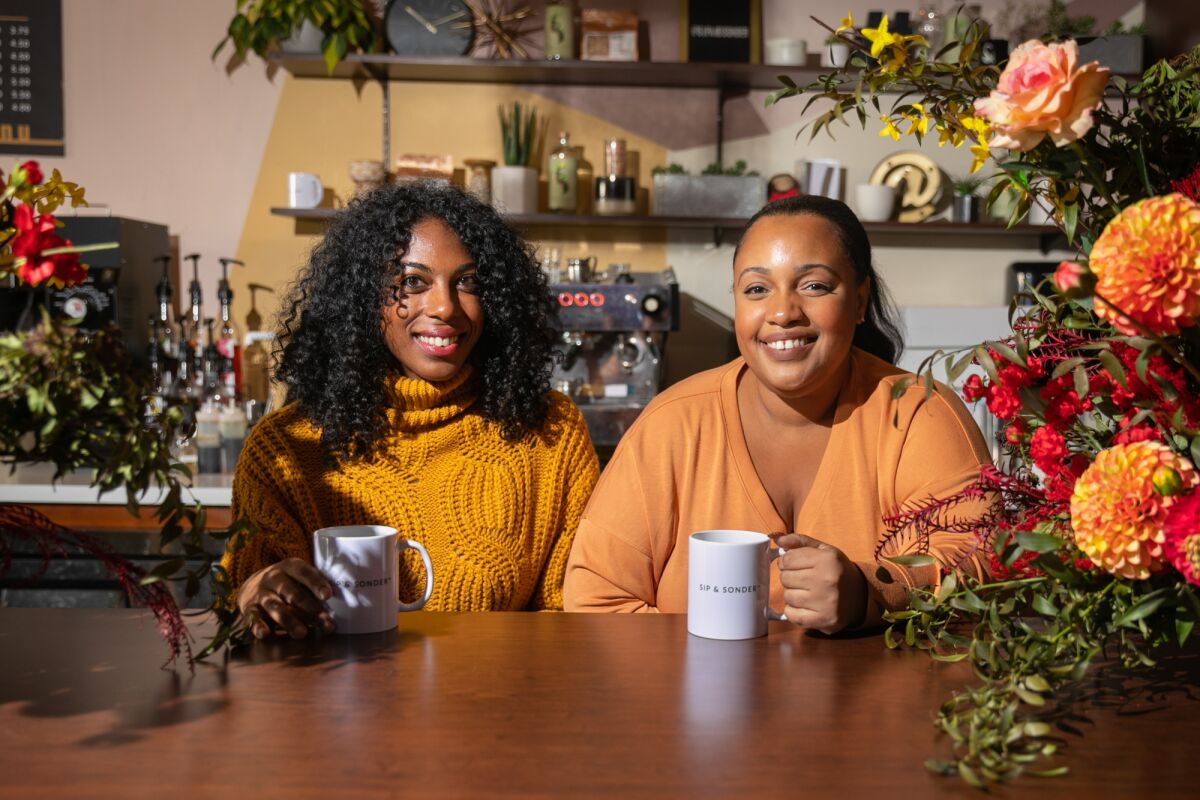  Two women pose for a portrait while holding white mugs.