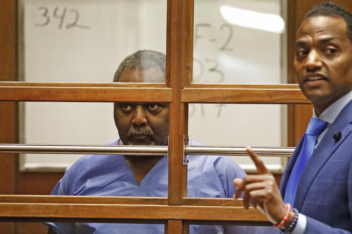 A man stands behind glass and bars in a courtroom as a lawyer speaks nearby