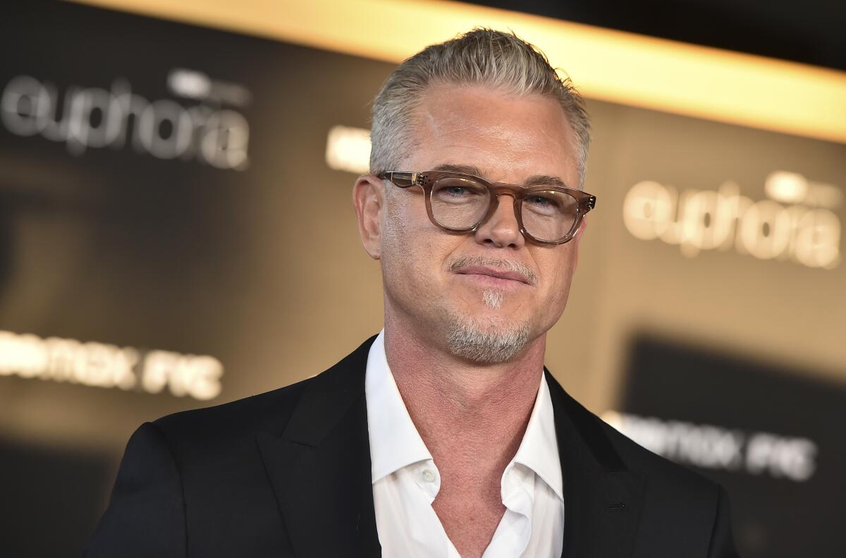 Eric Dane smiles very slightly in a black jacket, open-neck white shirt and tortoiseshell glasses