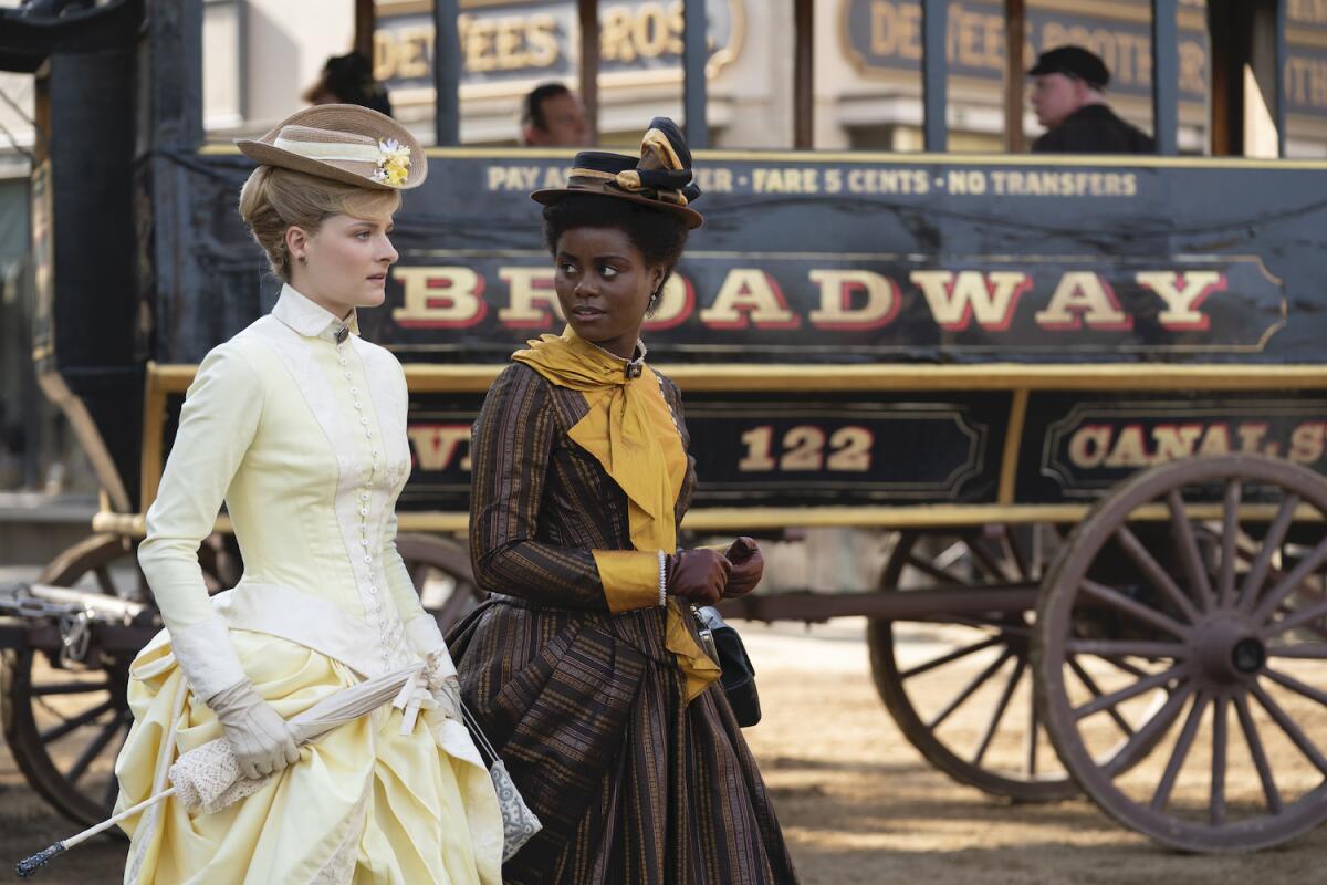 Two women wearing hats and late 19th century period dresses walk past a horse-drawn wagon labeled "Broadway."