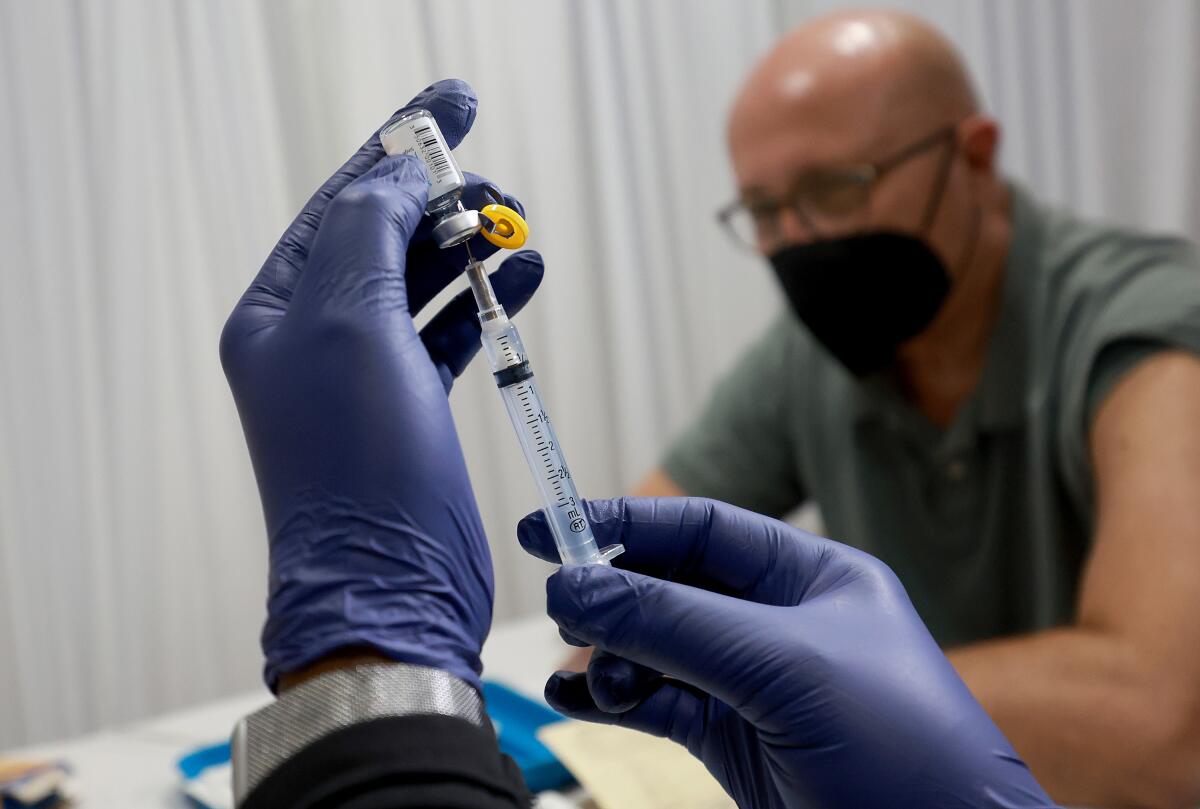 Gloved hands fill a syringe while a man sits in the background 