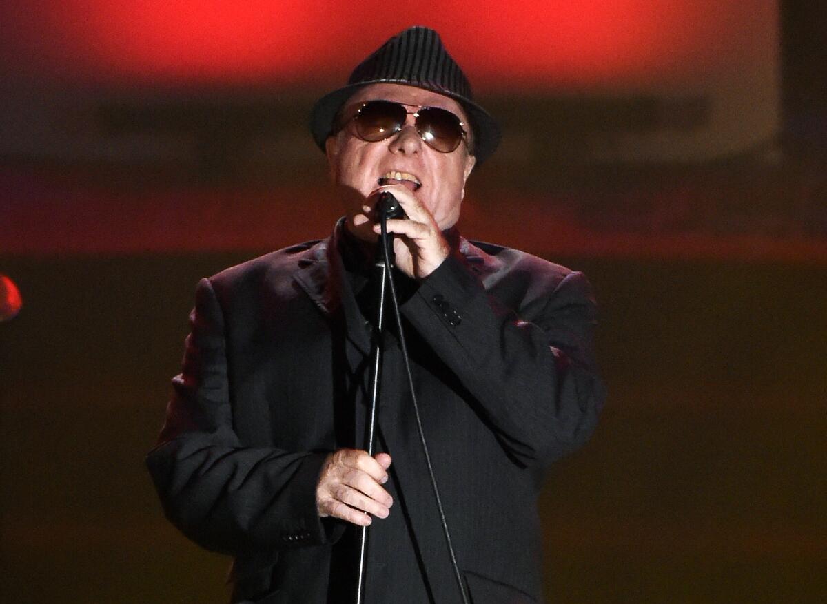 Van Morrison, wearing sunglasses and a hat, sings into a microphone.