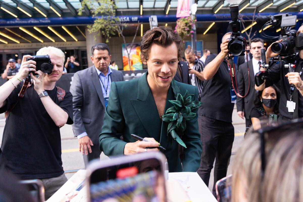 Harry Styles wears a green outfit and green flower as photographers take pictures behind him.
