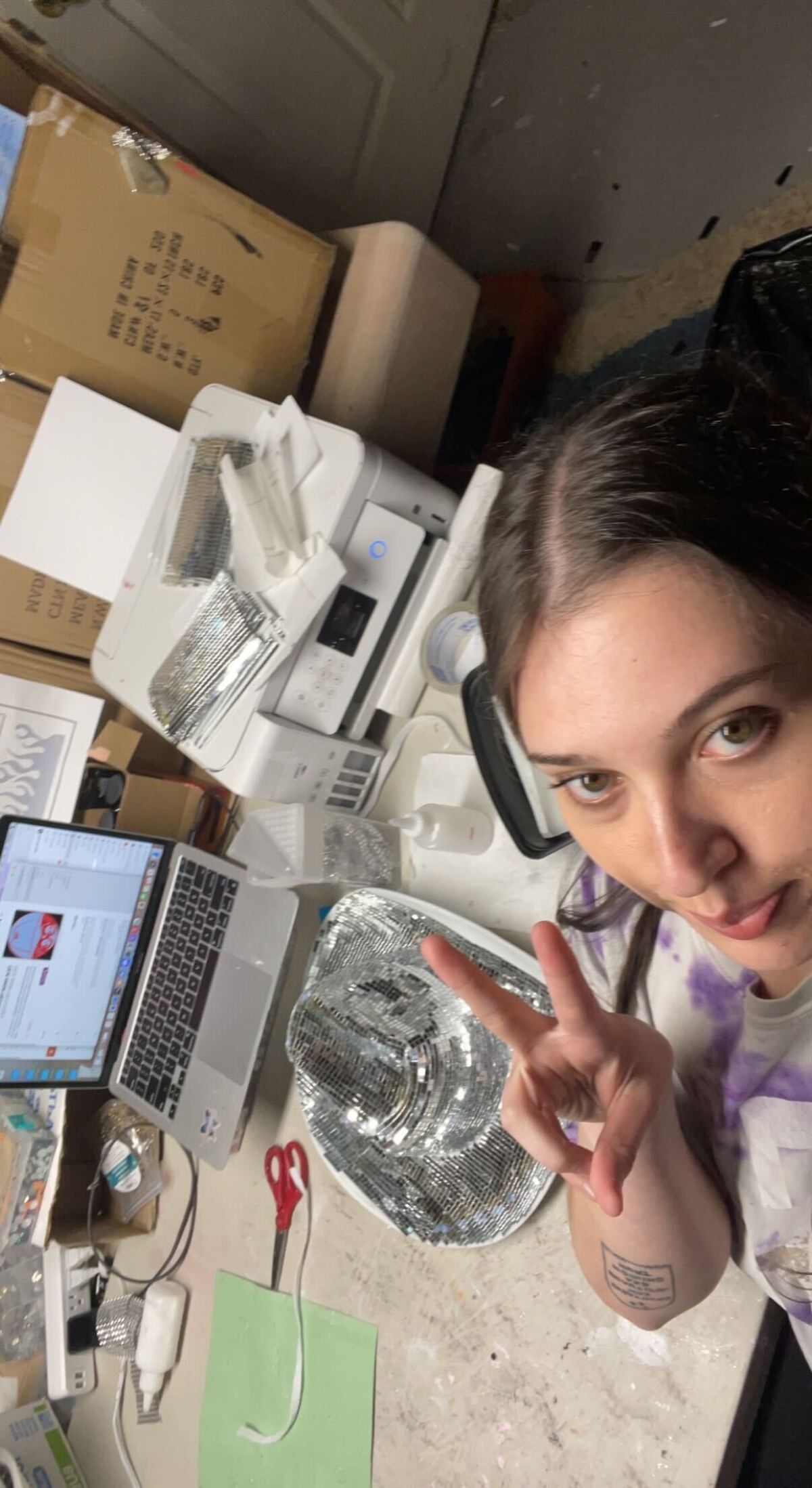 Woman holding up peace sign with cowboy hat and computer and crafts on a table