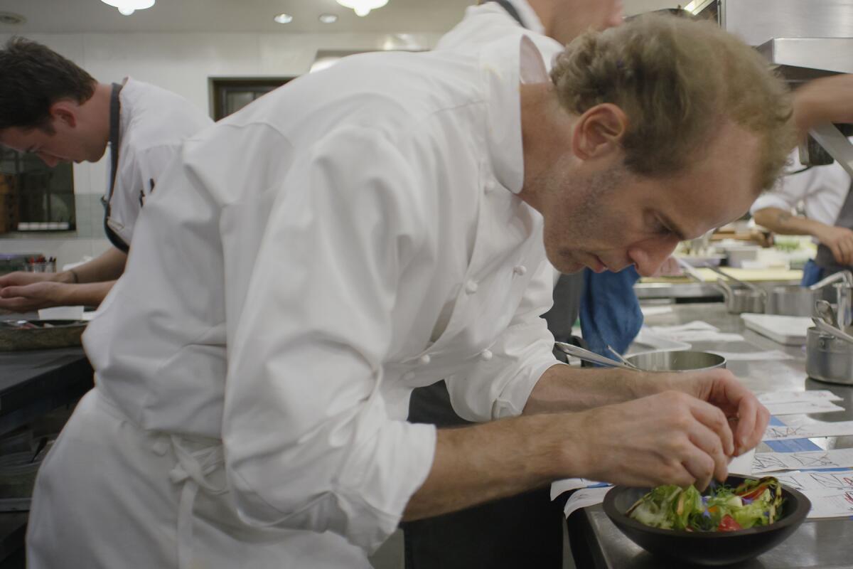 A chef leans over to fix a dish in a kitchen full of others doing the same.