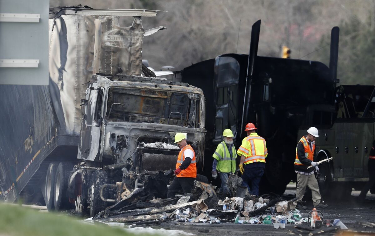 Workers clear debris from a road in front of a burnt semi-truck.