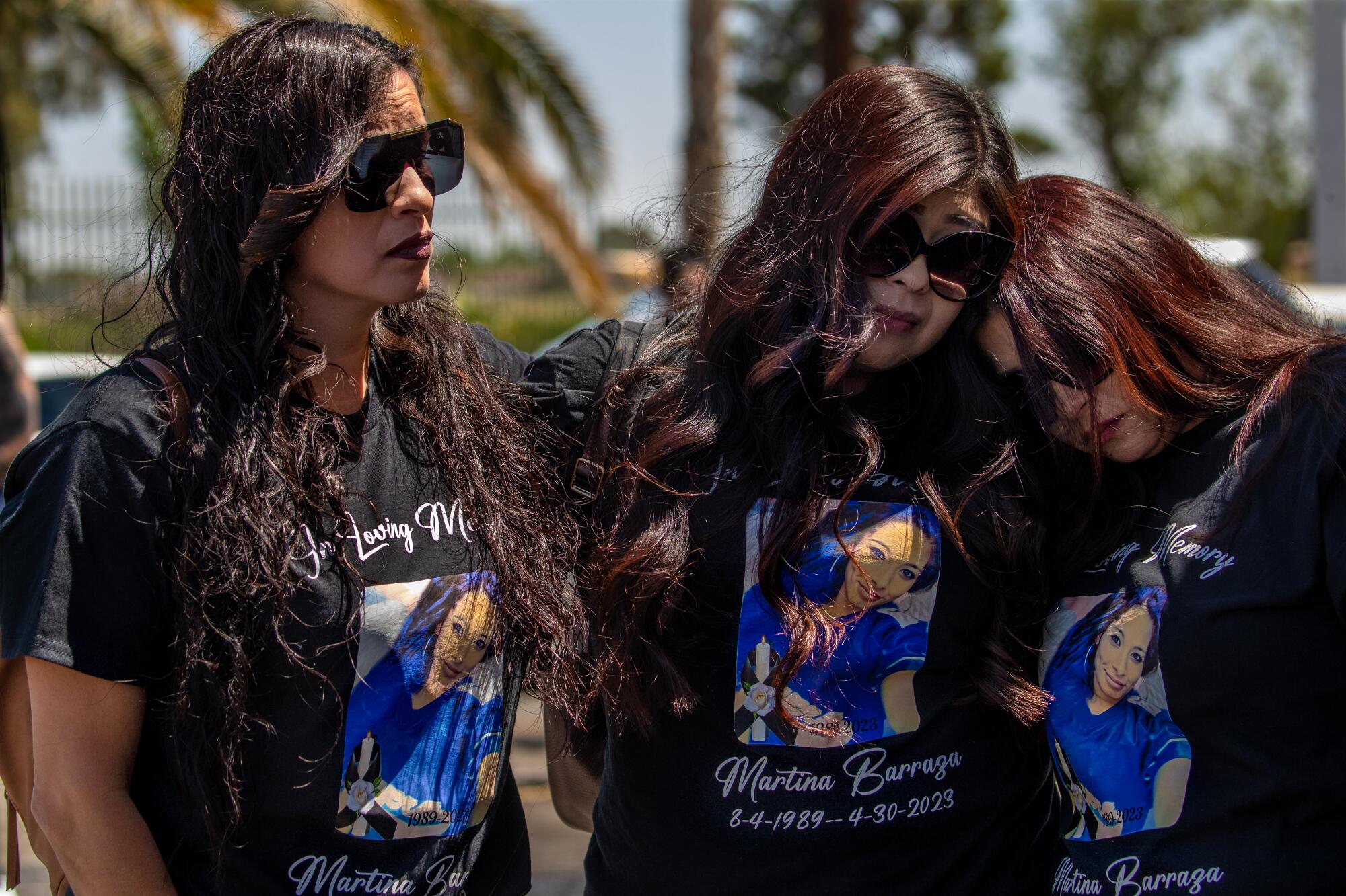 Three women wearing sunglasses and memorial T-shirts with a woman's photo embrace and lean on one another