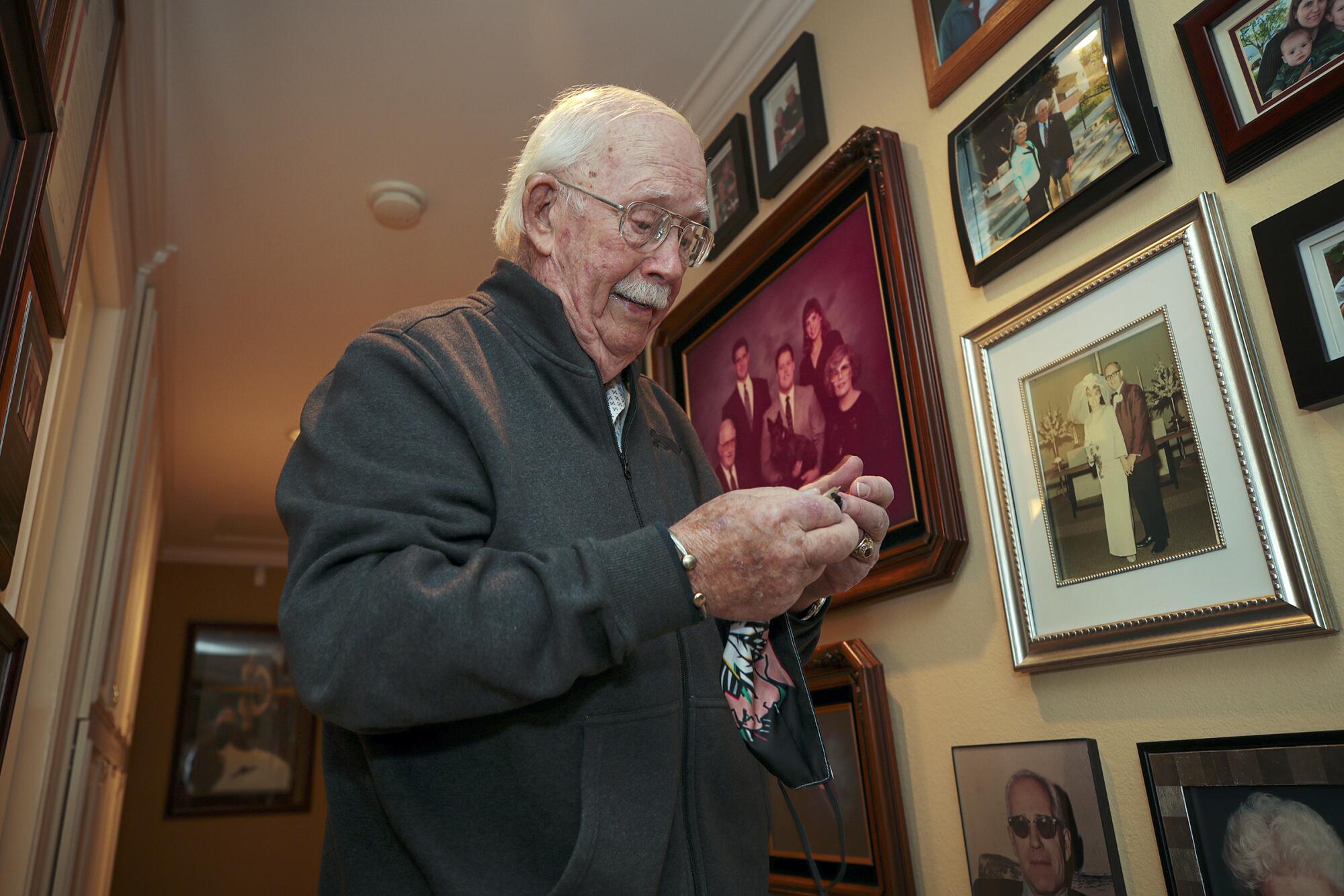 An elderly man looks at a handkerchief in his hands while standing next to a wall of family photos in his home