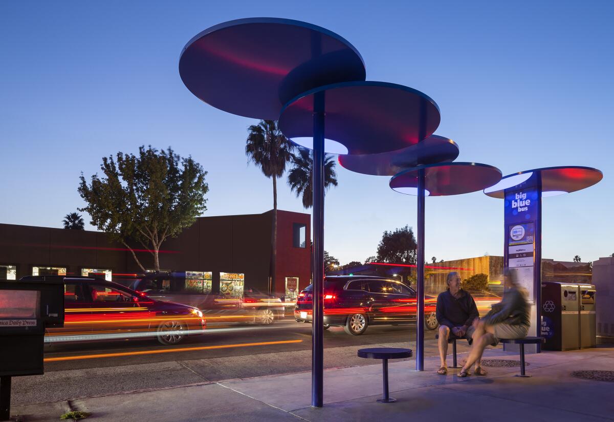 Two people sit under a shaded bus stop made of circular discs.