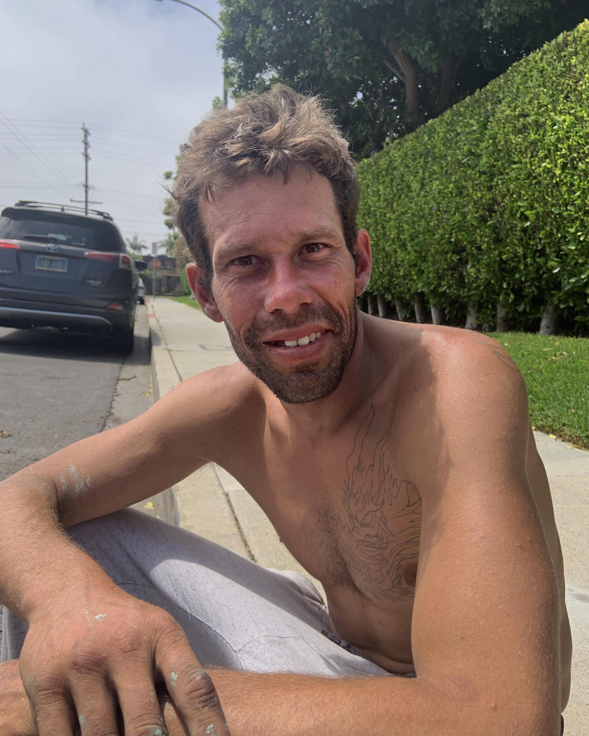 A vertical waist-up portrait of a shirtless man sitting on a curb on a sunny day, smiling and looking at the camera. 