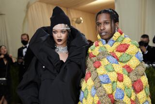 Rihanna in a puffy black coat and bedazzled headpiece standing next to ASAP Rocky in a colorful quilted oversized jacket