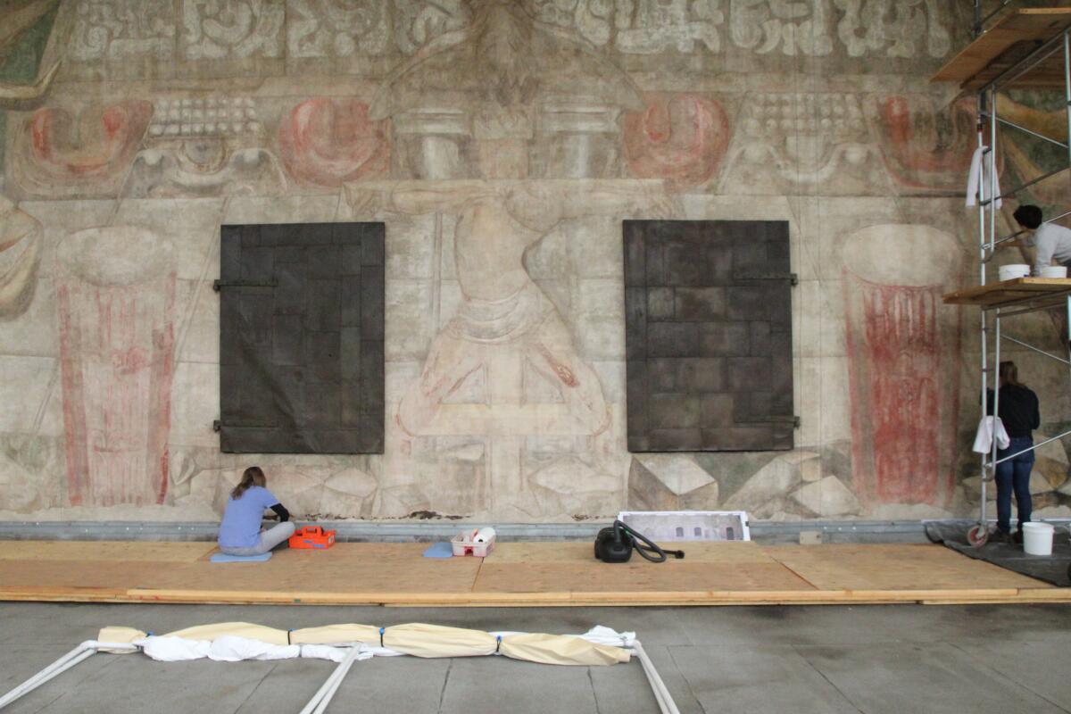 A worker in a blue shirt sits before a large, very faded mural in earth tones depicting vegetation and a crucified man