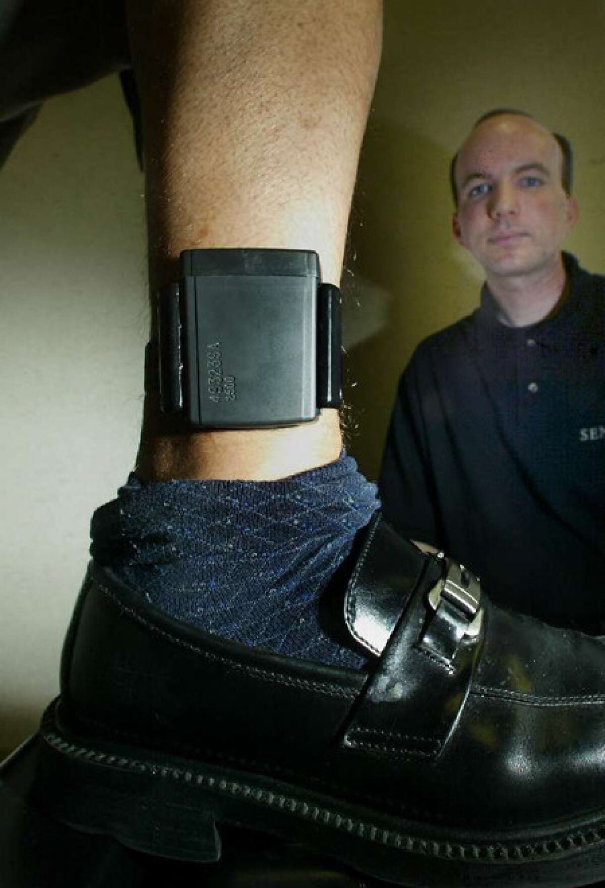 An electronic ankle style monitoring transmitter is demonstrated by a Sentinel Offender Services employee. In the background is Steve Hagstrom of Sentinel, who attached the device.