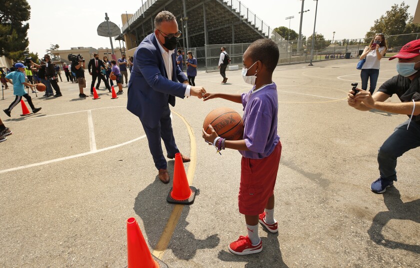 A man in a suit and face mask fist-bumps a child holding a basketball.