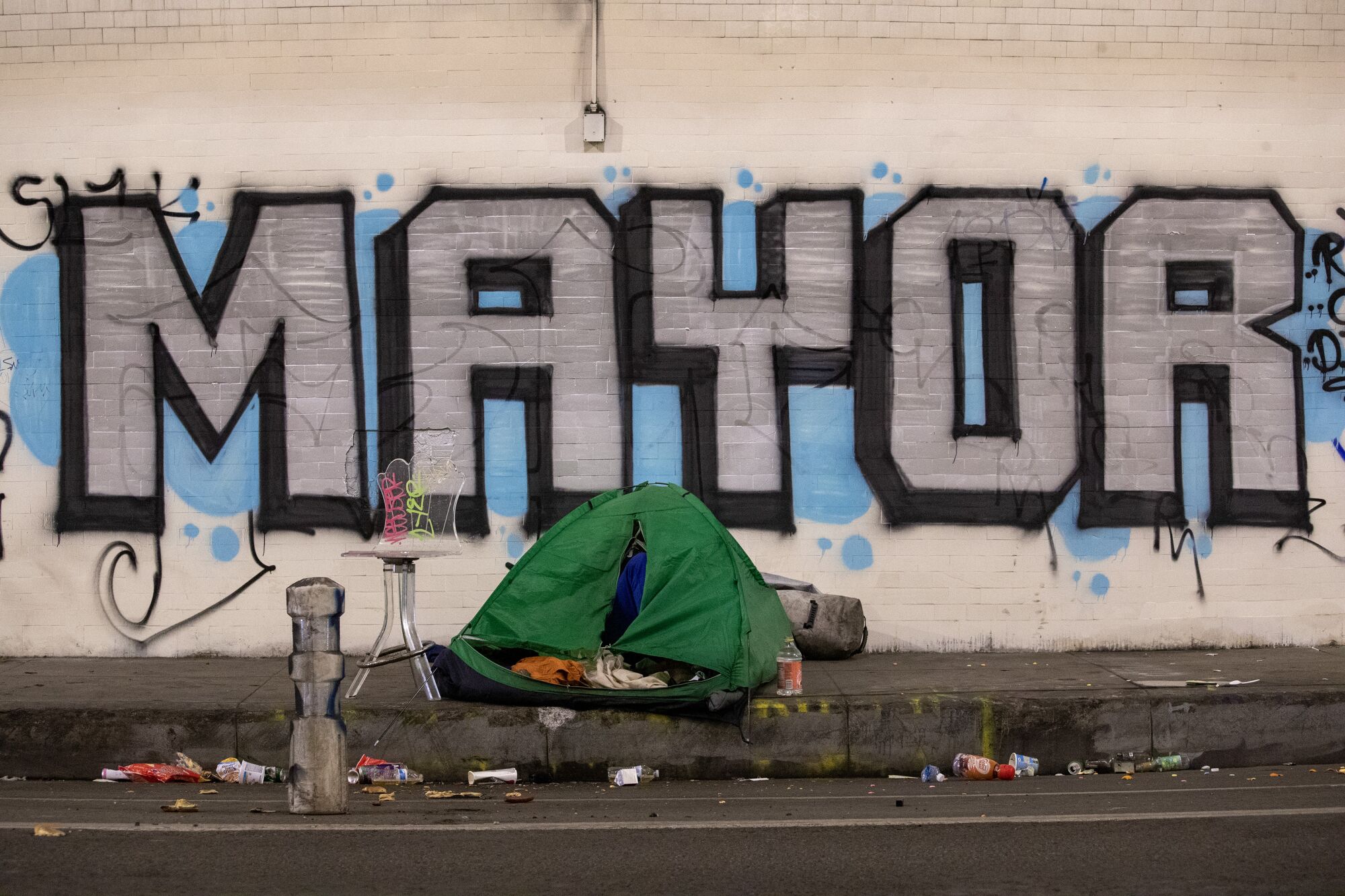 A homeless person sleeps in a tent on the sidewalk under graffiti that says "Mayor"