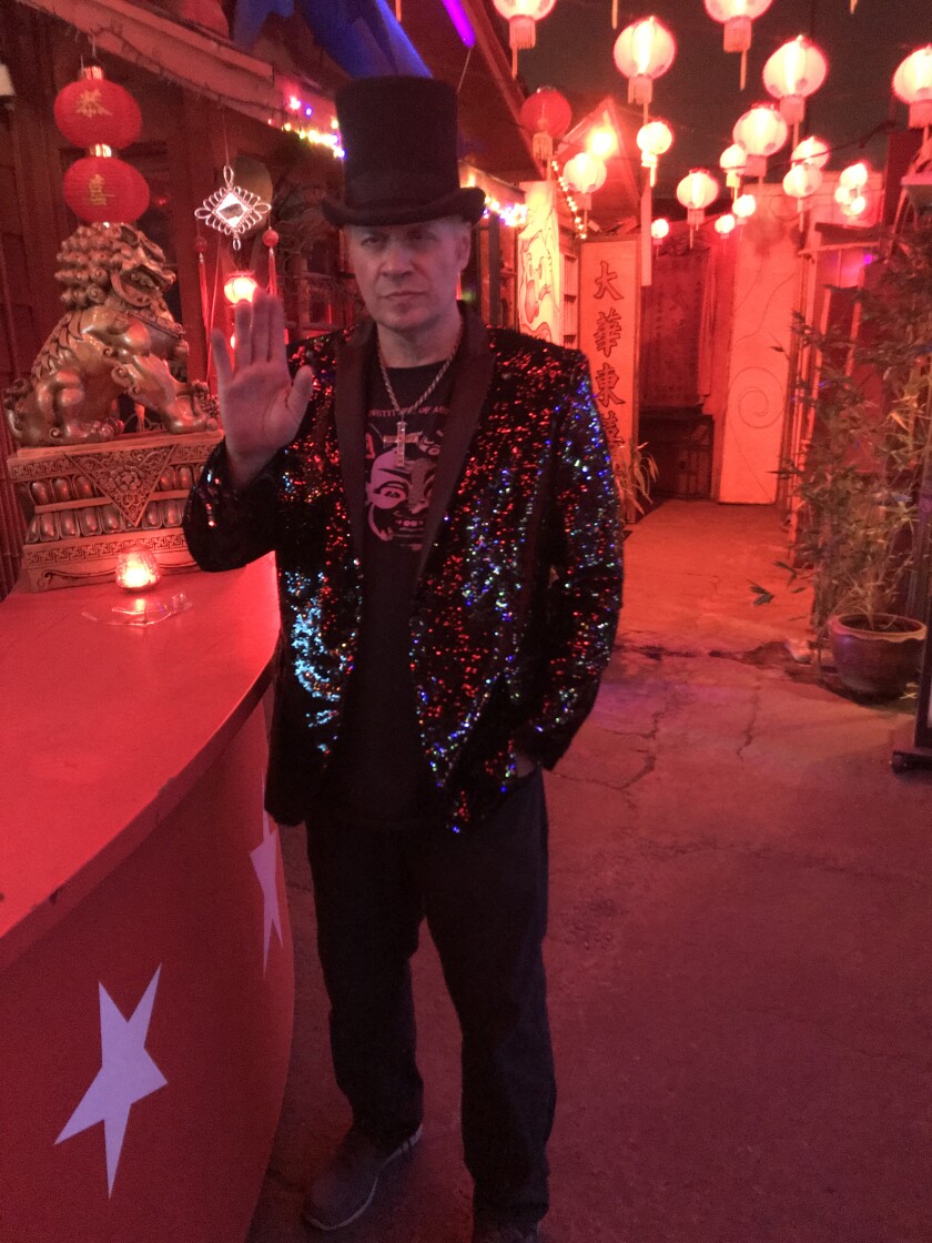 In a carnival environment, a man in a glittery top and jacket waves to the camera.