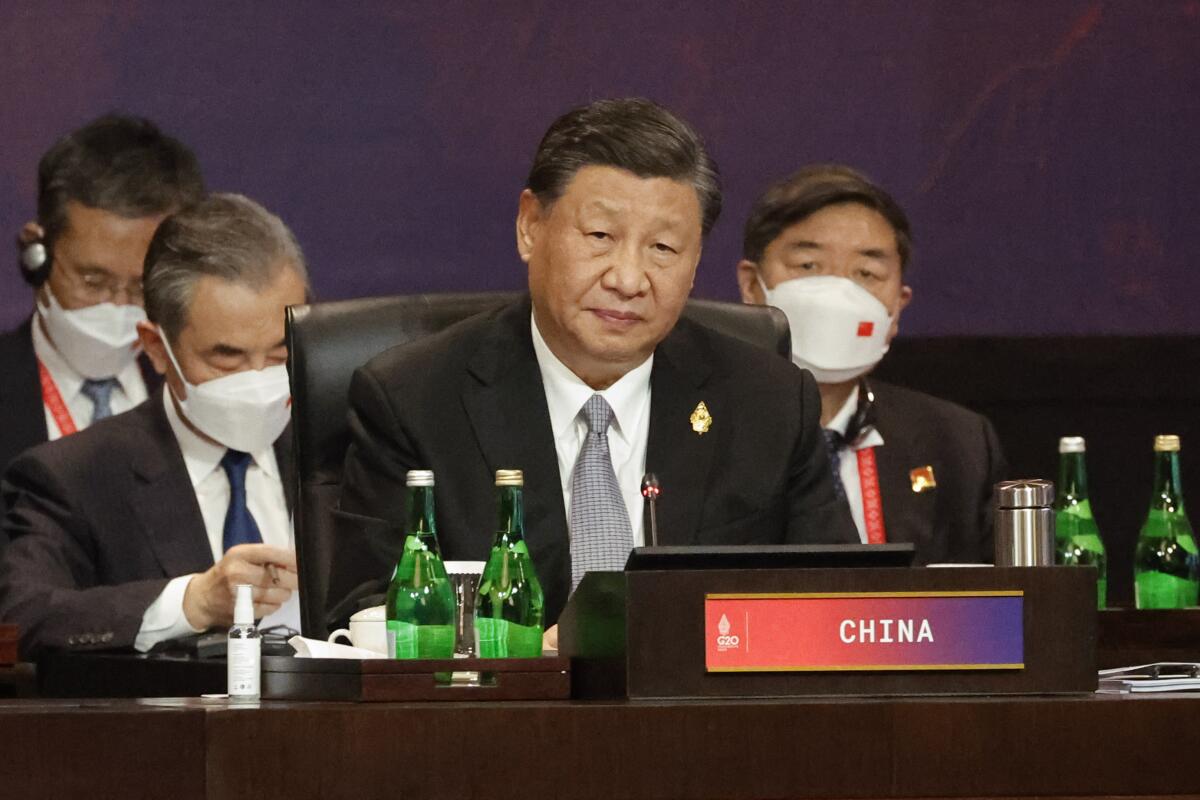 A man with dark hair, in a dark suit and gray tie, sits in front of other men in suits, behind a placard that says China