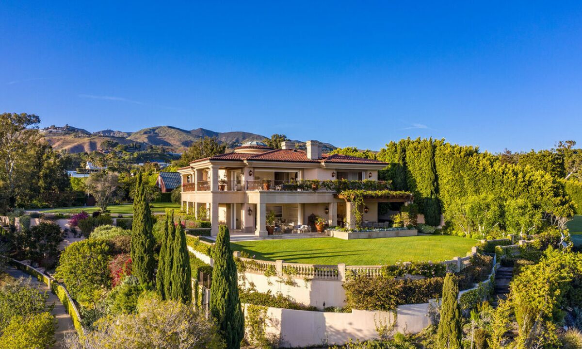 A mansion surrounded by lawn, bushes and trees, sitting in front of hills.