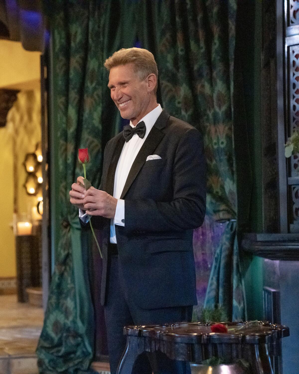 A smiling man in a tuxedo holds a red rose.