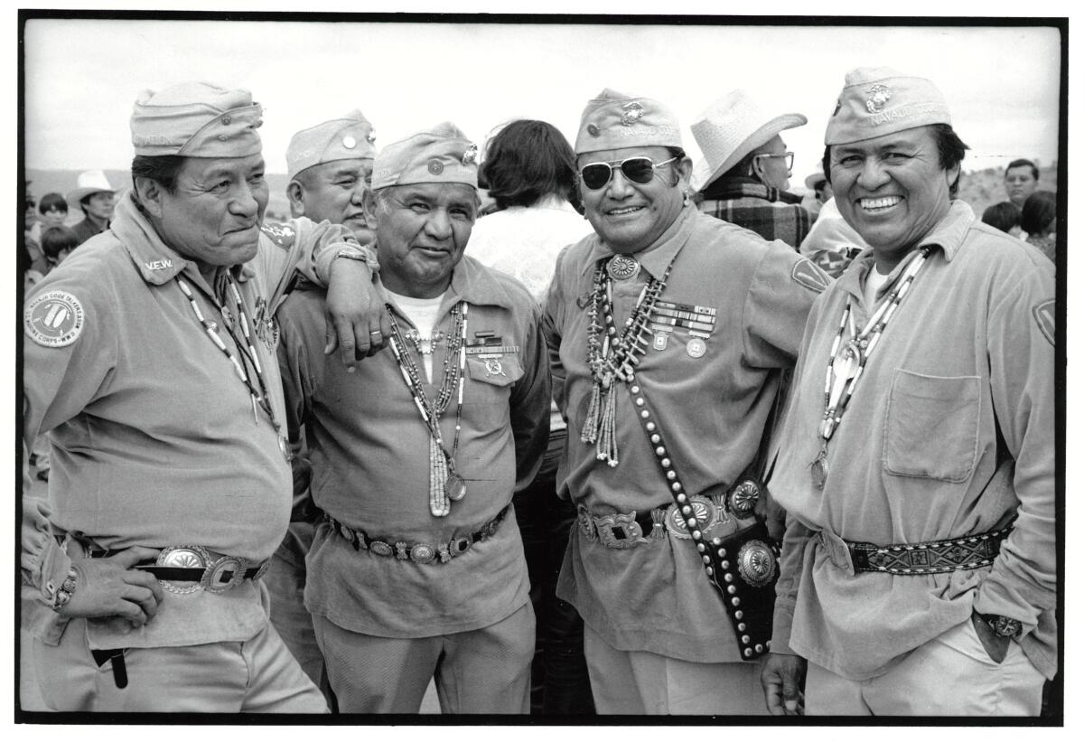 Four men in military uniform, with Native American necklaces and belts, pose for a picture near other people