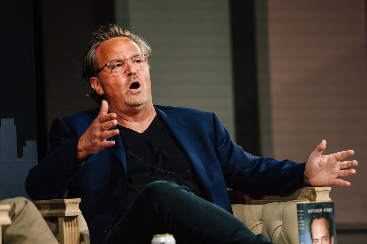 Matthew Perry sitting onstage, speaking with his hands outstretched
