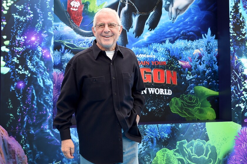 A smiling man in a black shirt stands in front of a poster for "How to Train Your Dragon: The Hidden World."