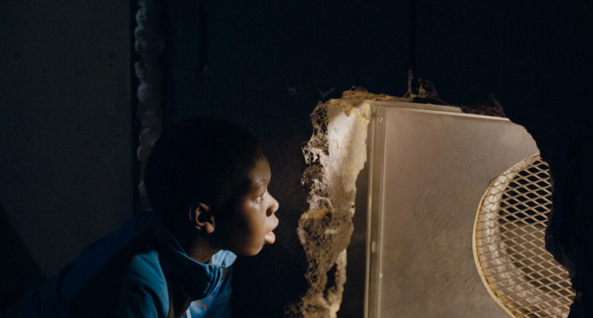 A boy in hiding peers through a screen in a wall.