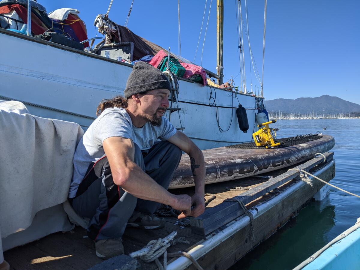 A man with a long hair and a beanie sits near a boat on the bay