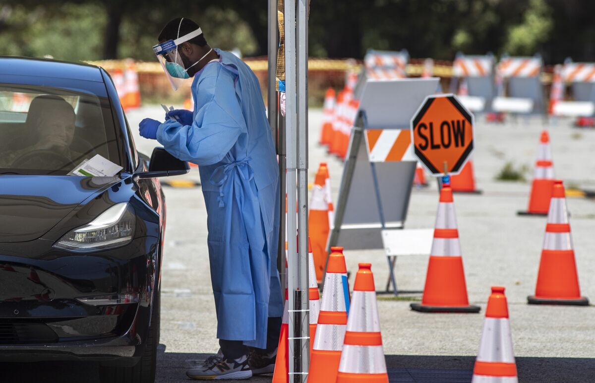 Surrounded by traffic cones, a man in gown, gloves, mask and face shield stands alongside a motorist.
