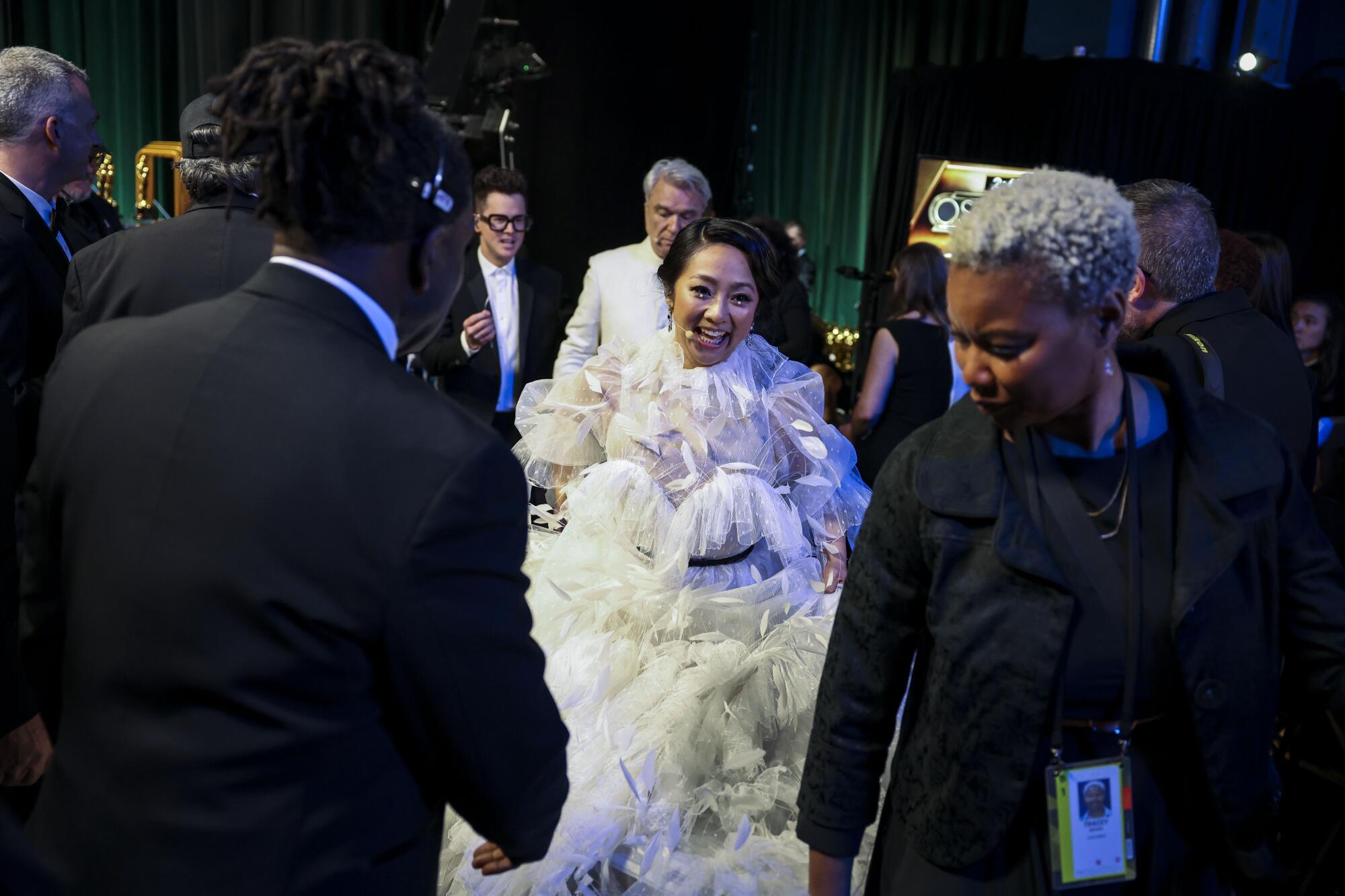 A woman in a flowy white dress and a man in a white suit smile and make their way through the backstage crowd.