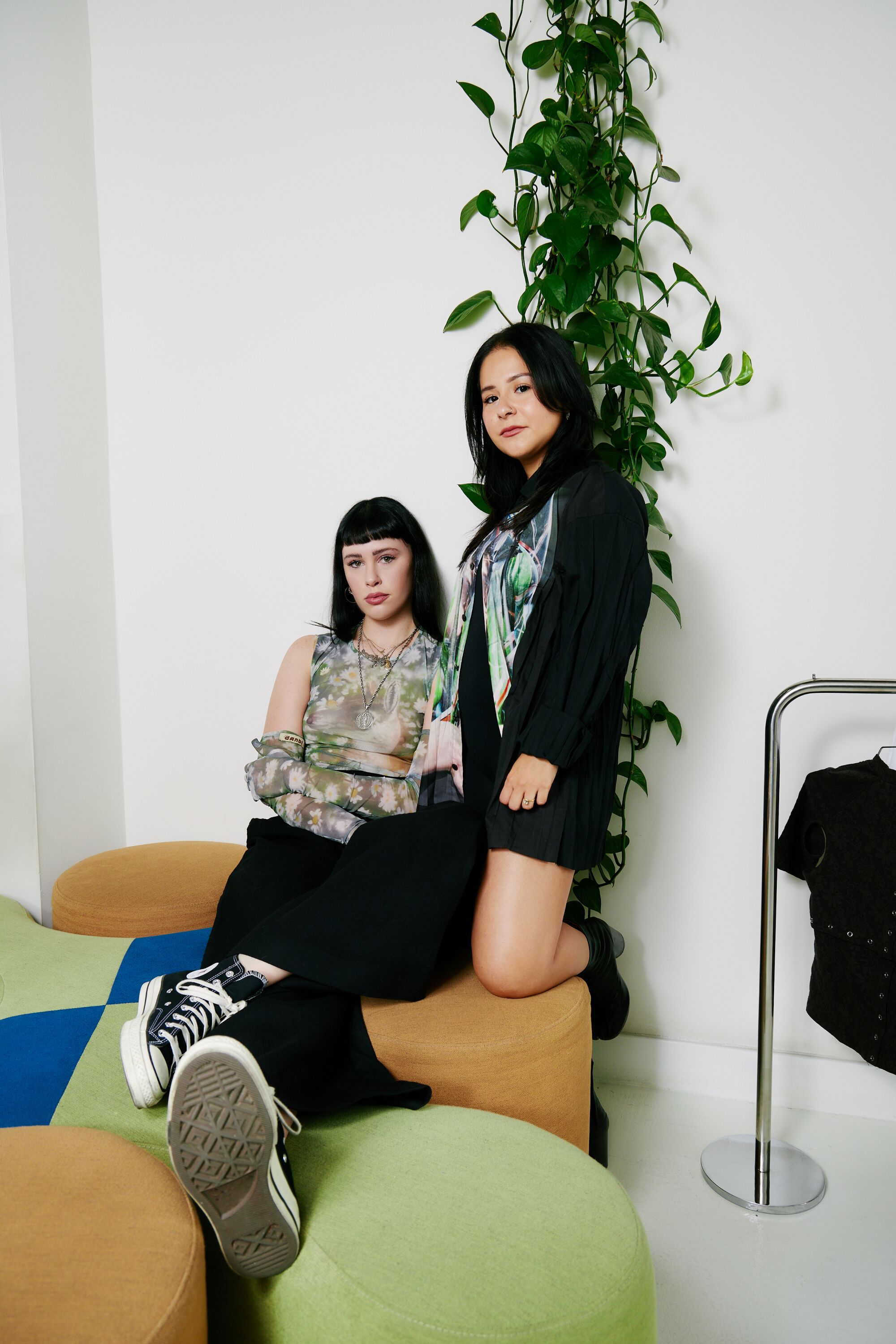 Two people among colorful ottomans, with vine-like greenery climbing the wall behind one.