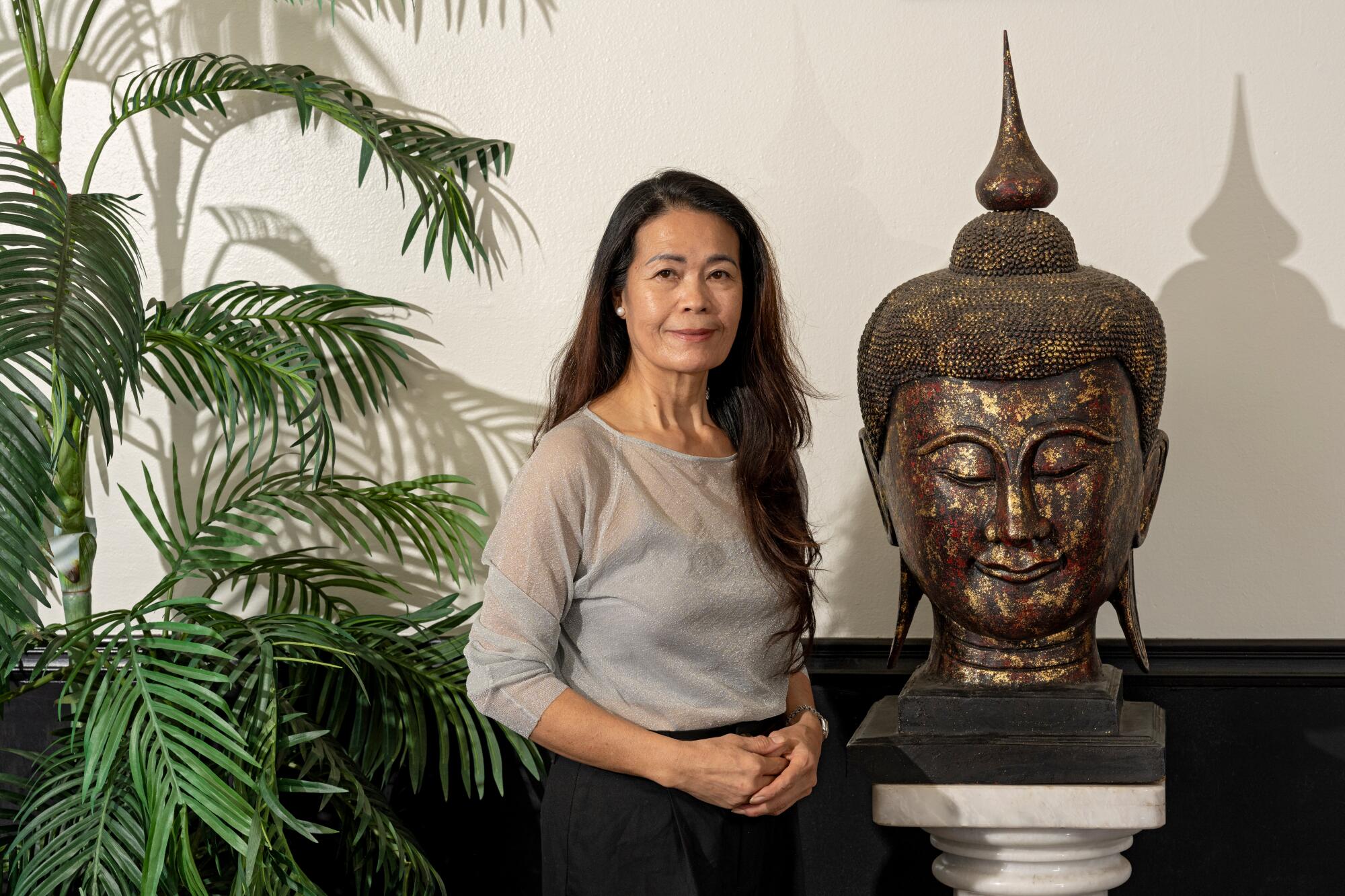 A woman stands next to a large metal Buddha head and a potted palm.