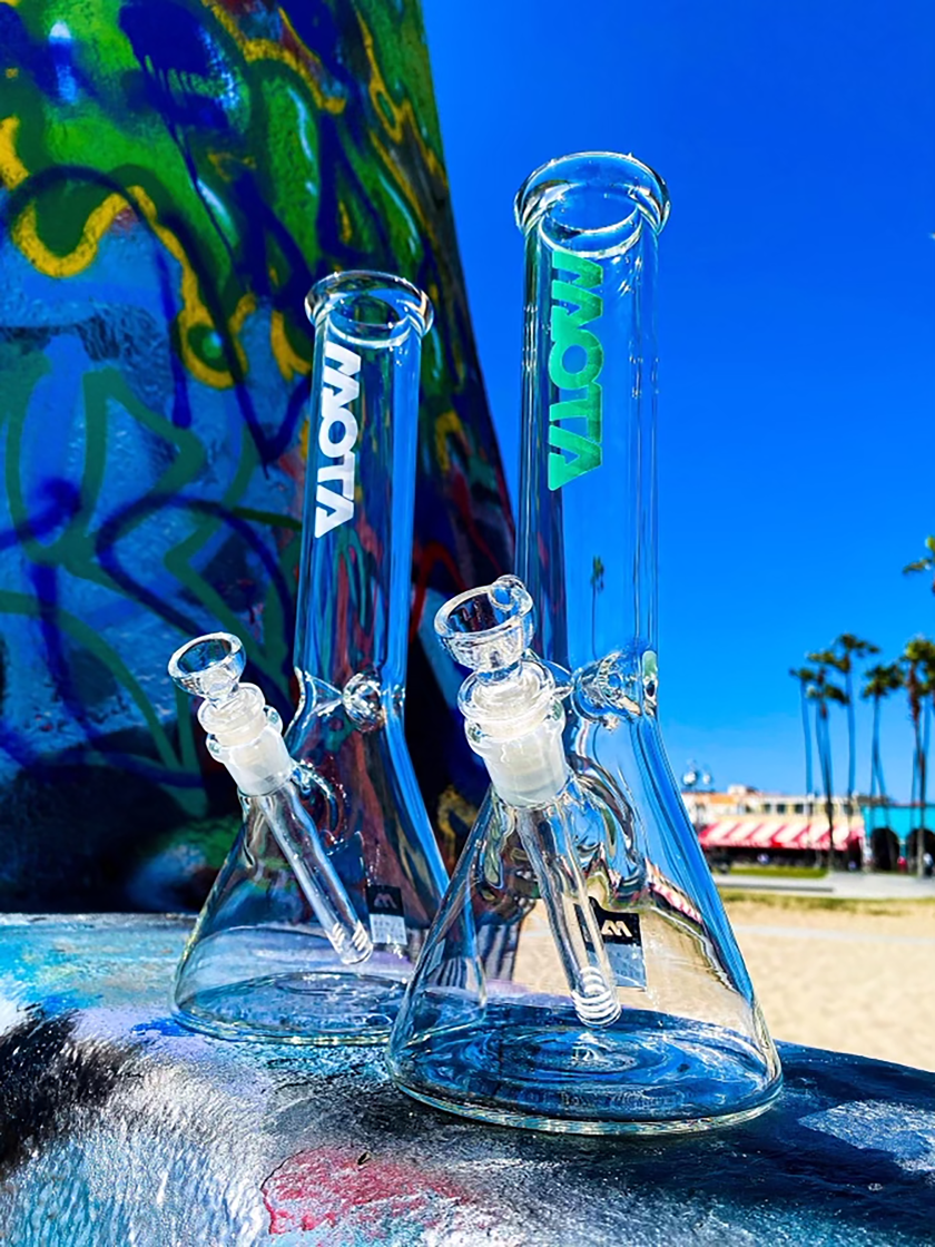 Two glass beaker bongs in the foreground with the beach and palm trees in the background.